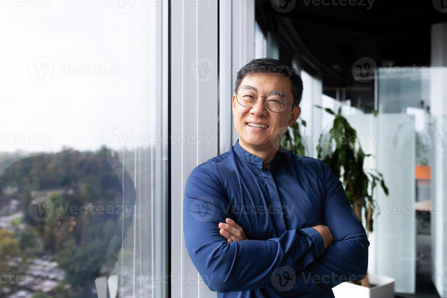 Portrait of successful asian boss inside office, businessman with crossed arms smiling and looking at camera, man in shirt and glasses happy with result of achievement at work. photo