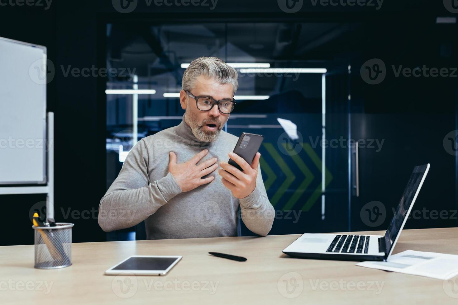 Mature businessman at work inside office, senior gray haired man shocked by bad news online reading from phone, boss upset over paper work holding smartphone. photo