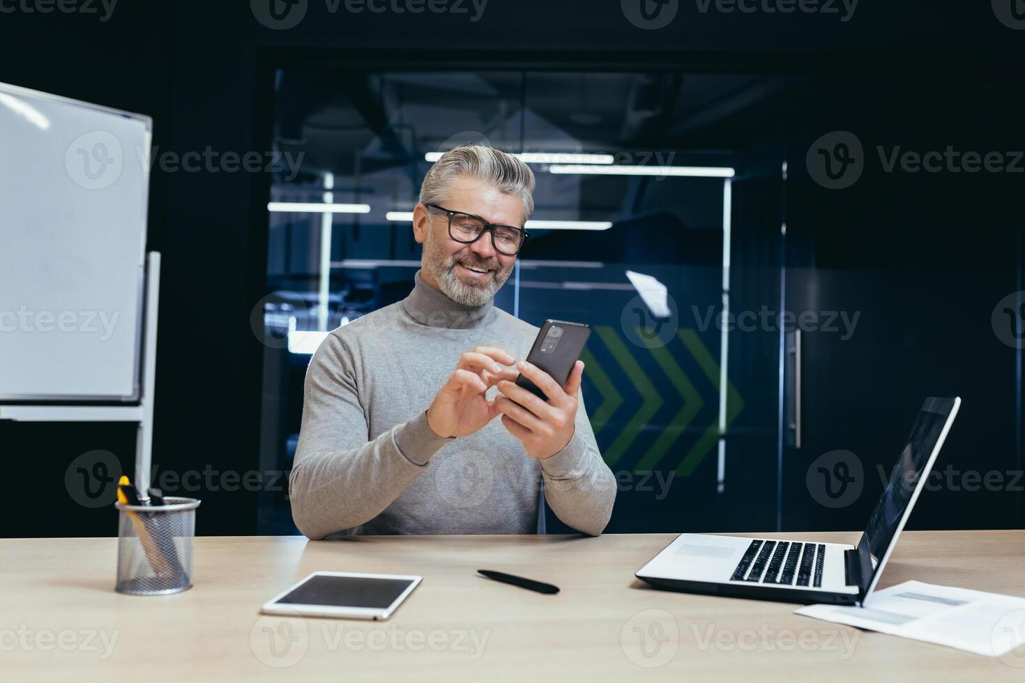 Mature successful businessman working inside office, senior gray-haired businessman using phone contentedly reading online message, man working with laptop at workplace, happy investor. photo