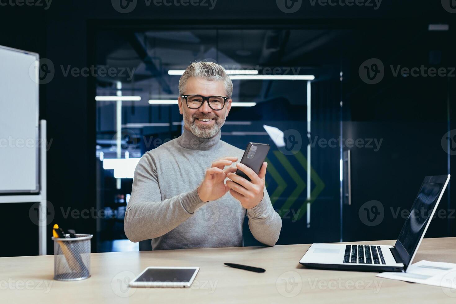 Portrait of successful mature investor inside office with work, gray-haired businessman smiling and looking at camera, man holding phone, typing message, using laptop at work. photo