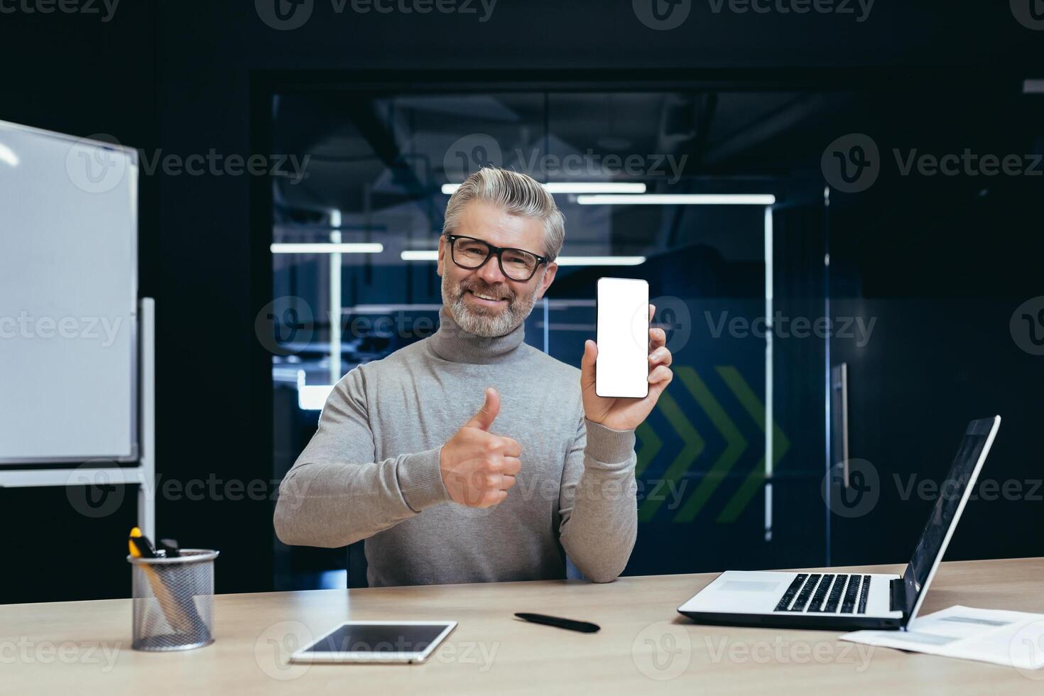 Portrait of mature businessman inside office, senior boss smiling looking at camera and showing white blank smartphone screen, man holding affirmative and recommending thumbs up working with laptop. photo
