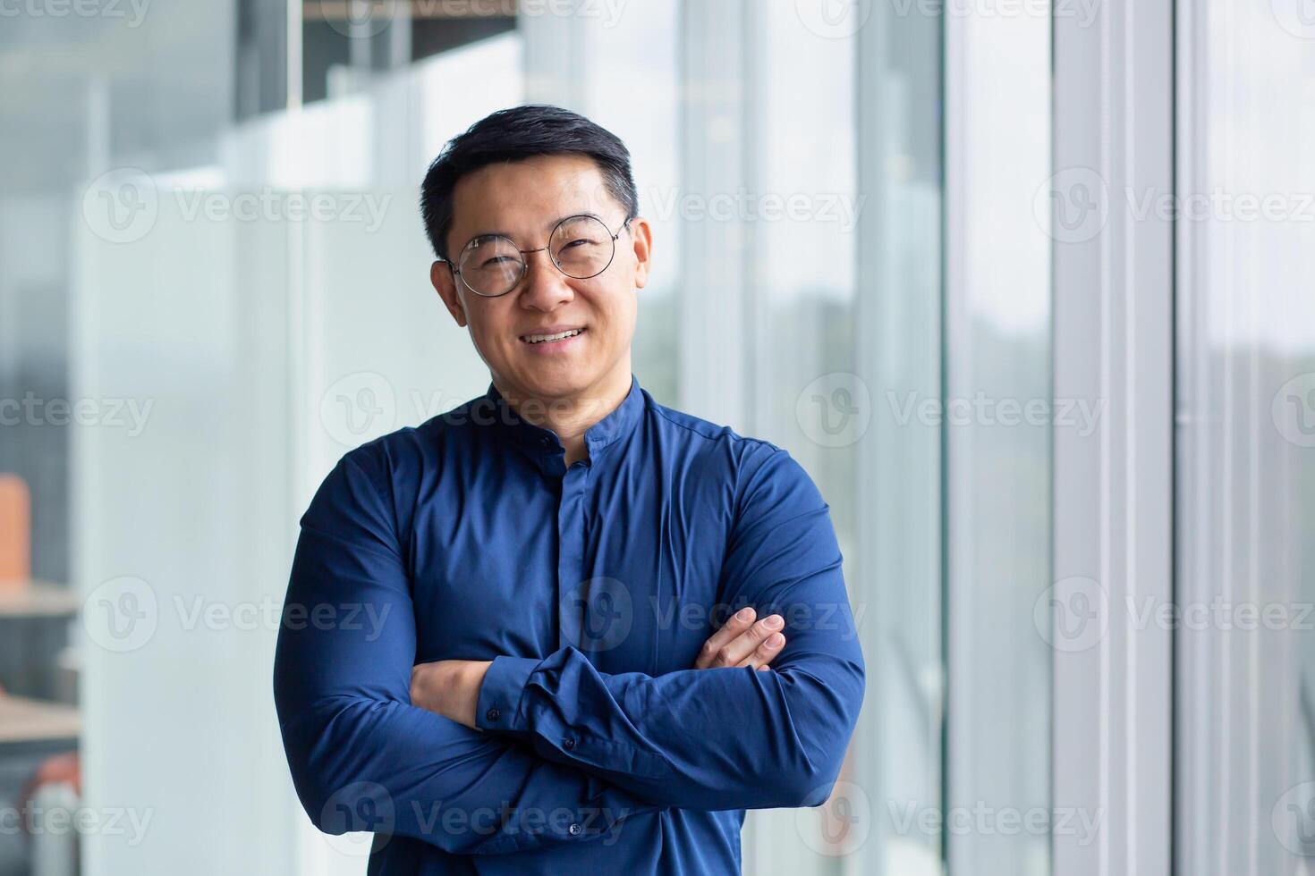 Portrait of successful mature boss, senior businessman in glasses Asian looking at camera and smiling, man with crossed arms working inside modern office building. photo