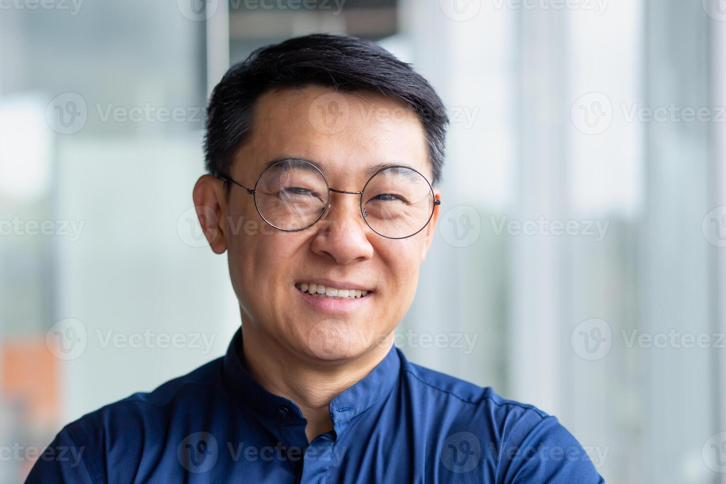 Close up photo portrait of successful and happy Asian businessman, mature boss with glasses working inside modern office building senior investor smiling and looking at camera