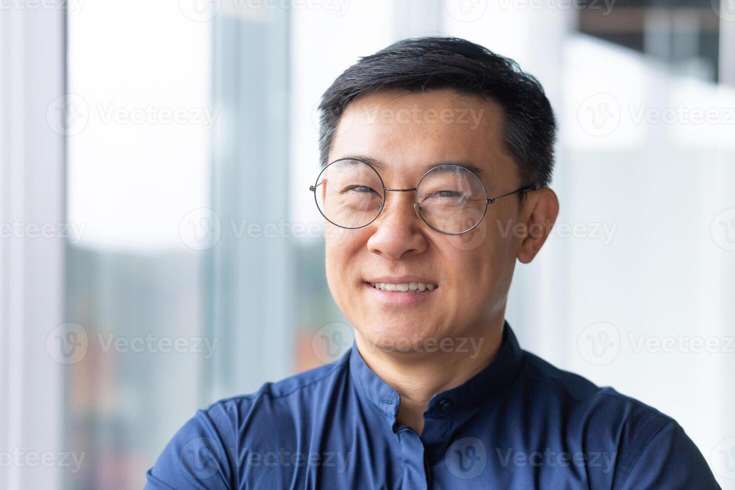 Close-up portrait of smiling asian man inside office, man smiling and looking at camera in glasses and shirt, successful engineer programmer engineer developer in development company at work. photo