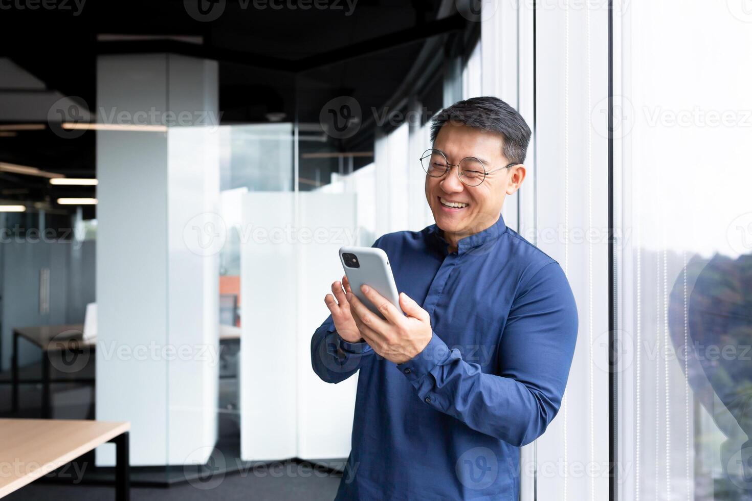Asian businessman inside the office in a casual shirt and glasses is using the phone, the boss is holding a smartphone in his hands reading news and typing a message smiling. photo