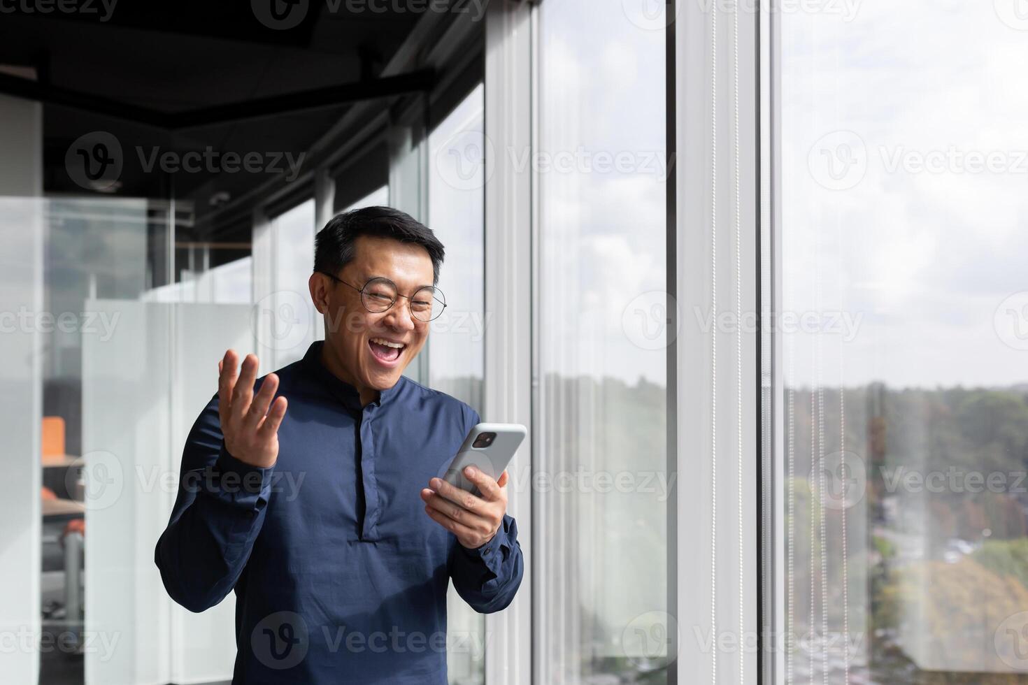 Asian man using phone standing near window inside office building, programmer in shirt and glasses celebrating victory and successfully completed software development, reading news from smartphone. photo