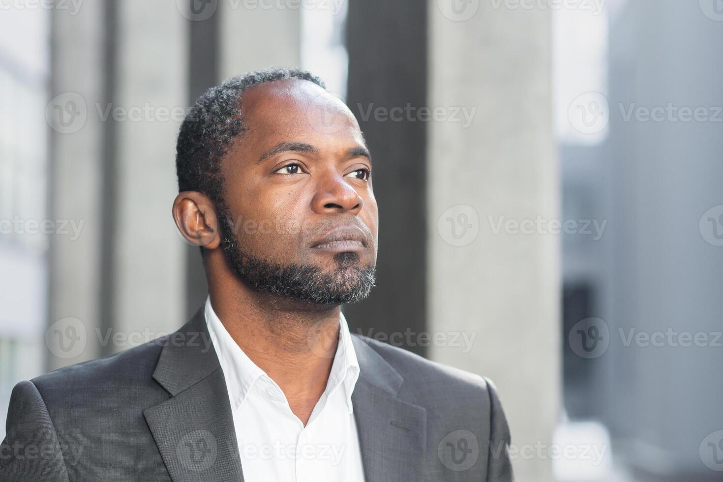 Close-up photo. Portrait of a successful African American businessman, leader, boss. He is standing in a suit near the office center, looking to the side with a confident look. photo