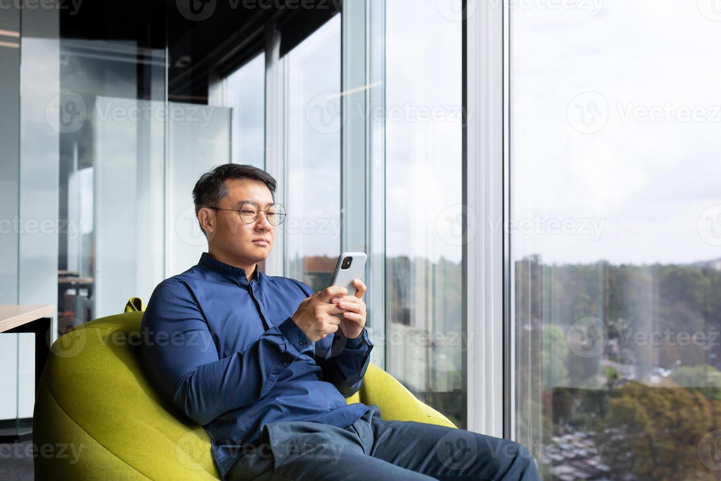 Serious thinking asian man reading news on phone, programmer using app on smartphone testing new program, businessman worker sitting on soft chair near window in modern office interior. photo