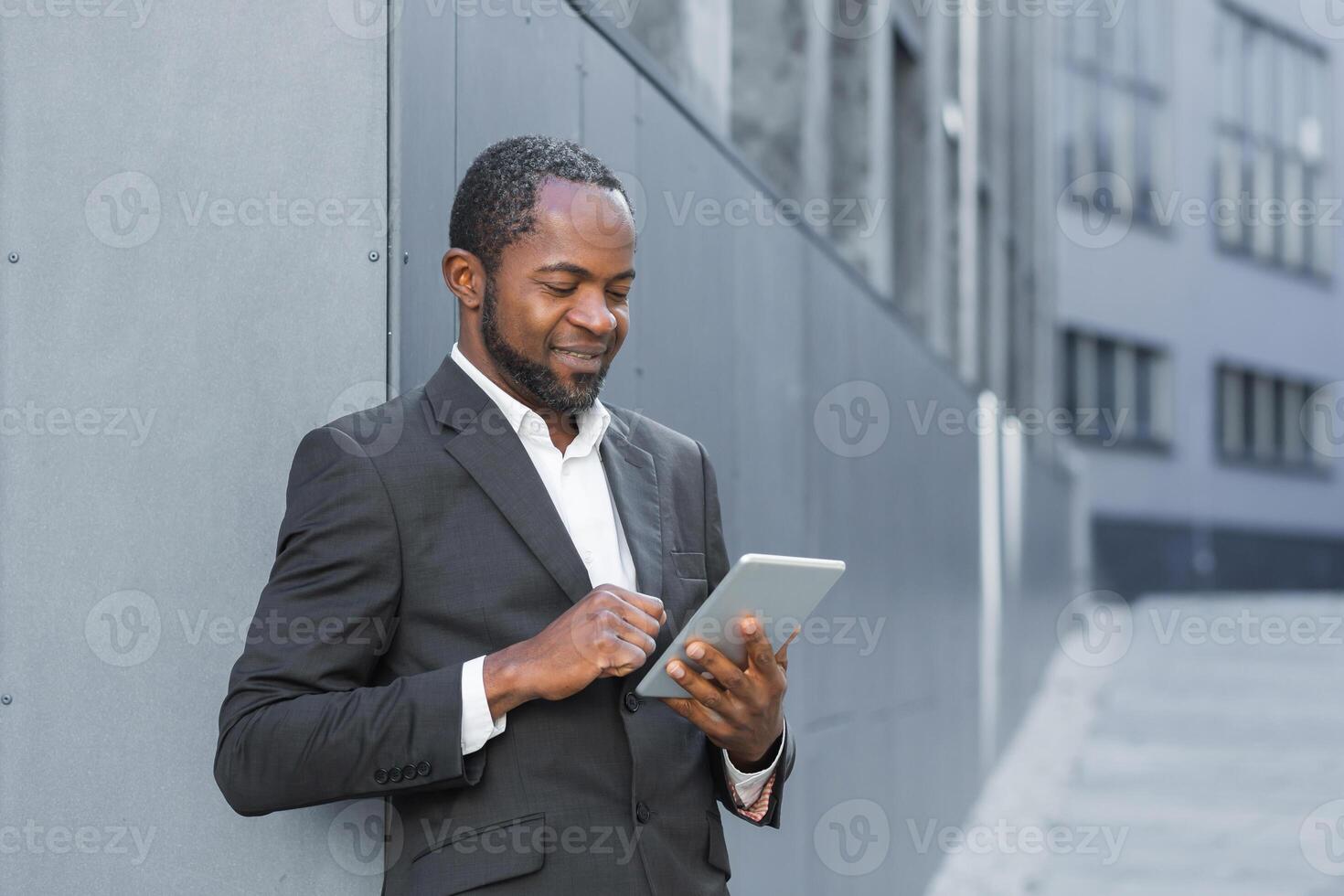 Photo from below. An African American businessman is standing outside an office center in a suit and using a tablet. Dials, communicates, smiles.
