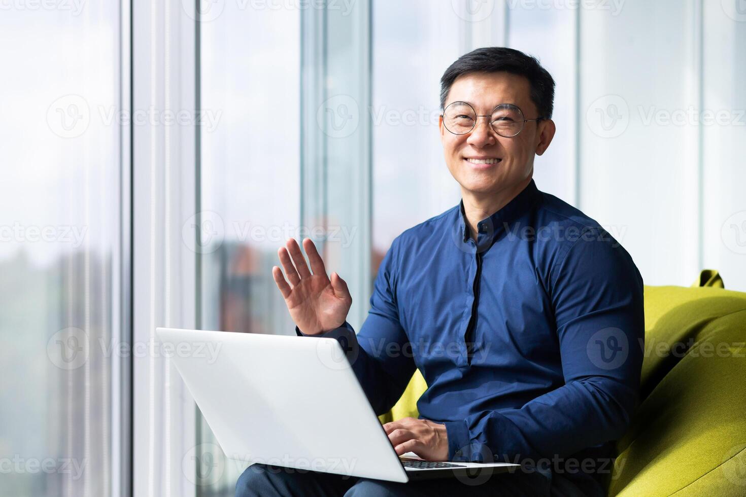 Portrait of successful smiling asian man inside office, man smiling and looking at camera near window, businessman working sitting with computer, waving hand in greeting gesture. photo