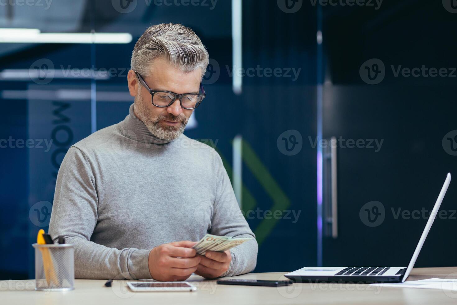 Serious mature investor working inside office, senior gray haired businessman counting money cash dollars, businessman working inside contemporary office using laptop at work. photo