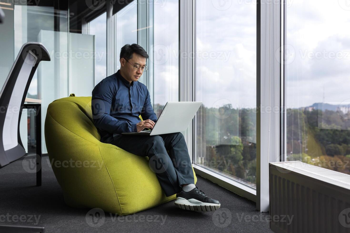 Asian young office worker sitting on soft ottoman chair, programmer using laptop for writing code and programming testing new program, man sitting near window inside office. photo