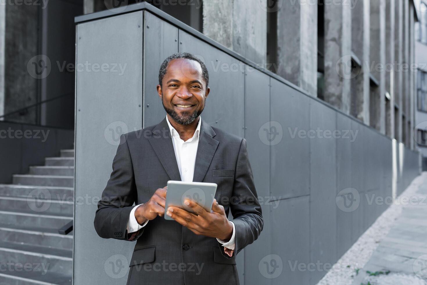 Portrait of a successful African American boss, a businessman in a suit smiling and looking at the camera, a man holding a tablet computer in his hands, a successful investor browsing the Internet. photo