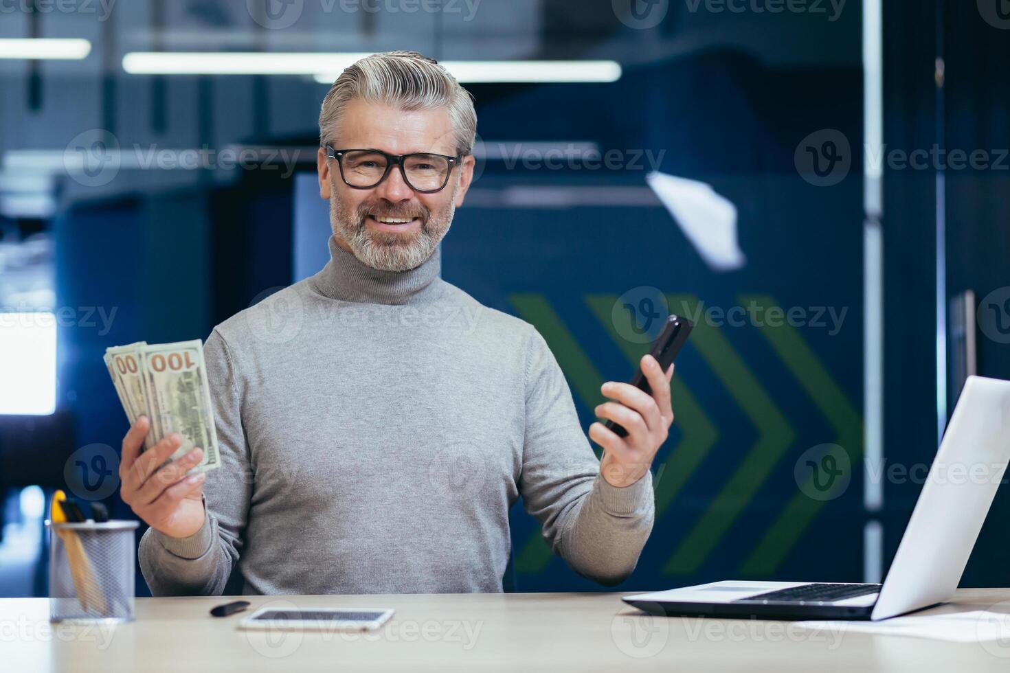 Portrait of mature successful businessman inside office, senior gray haired man smiling and looking at camera, investor holding phone and money cash dollars in hands, banks happy with income online. photo