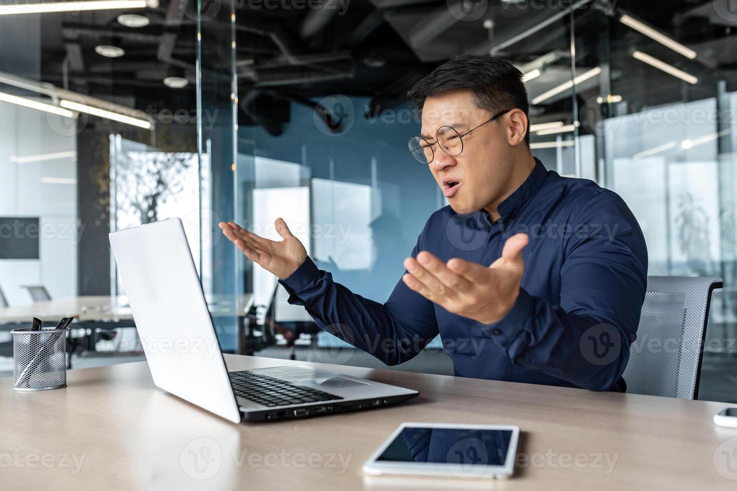 Frustrated and angry asian boss yelling at laptop, failure man working inside twisted office building at desk. photo