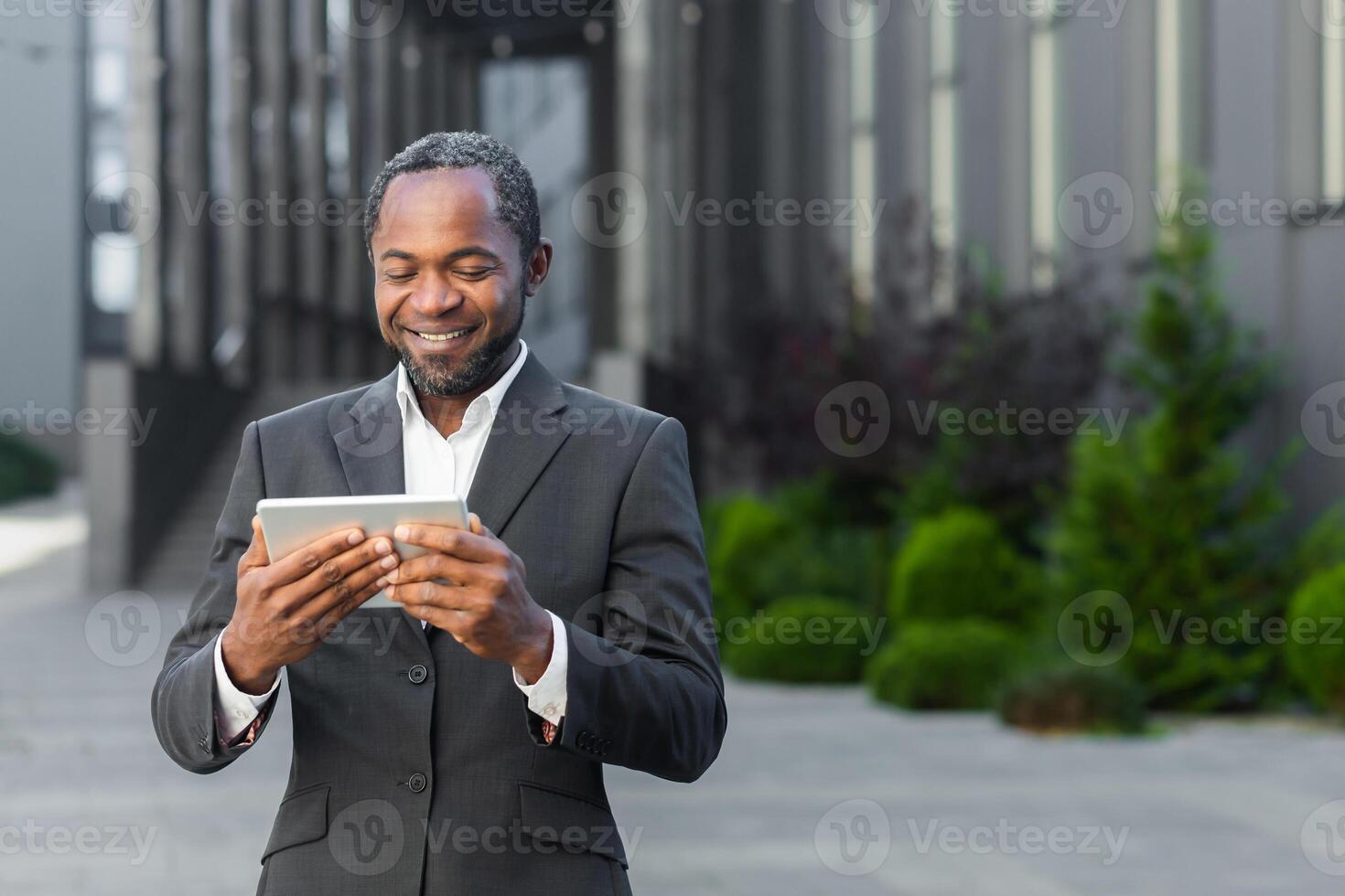 African american man in business suit outside office building watching online with using app on tablet computer, businessman smiling and happy. photo