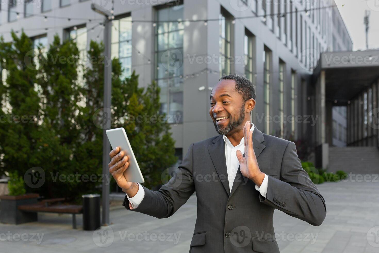 An African-American businessman is standing outside in a suit, holding a tablet and talking on a call. photo