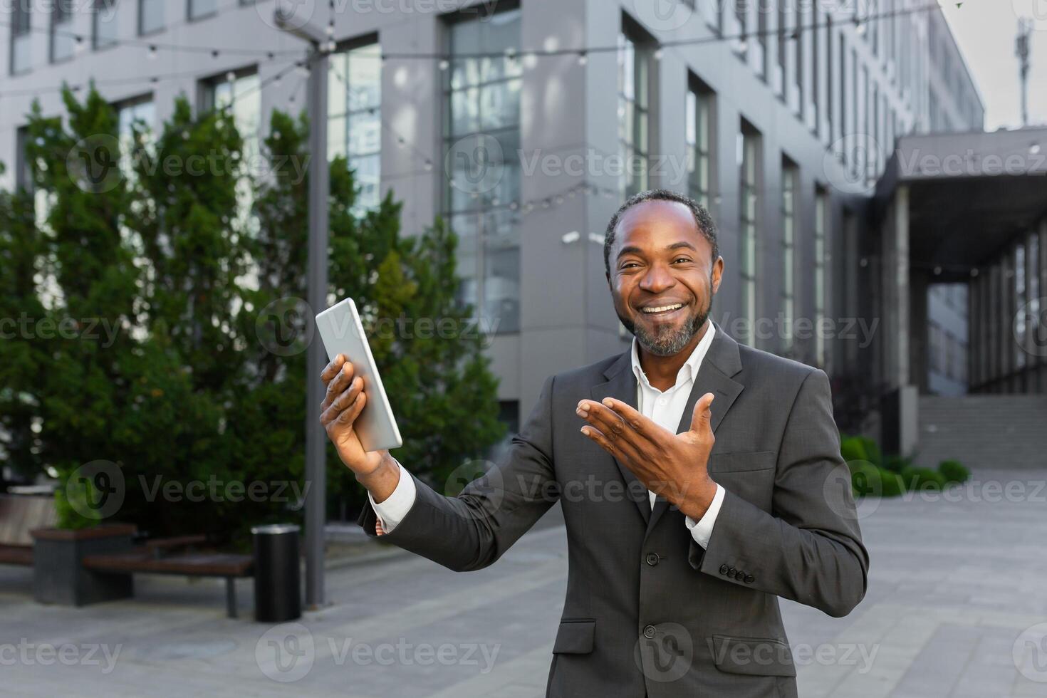 An African-American businessman is standing outside in a suit, holding a tablet and talking on a call. He looks at the camera, points to the gadget, smiles. photo