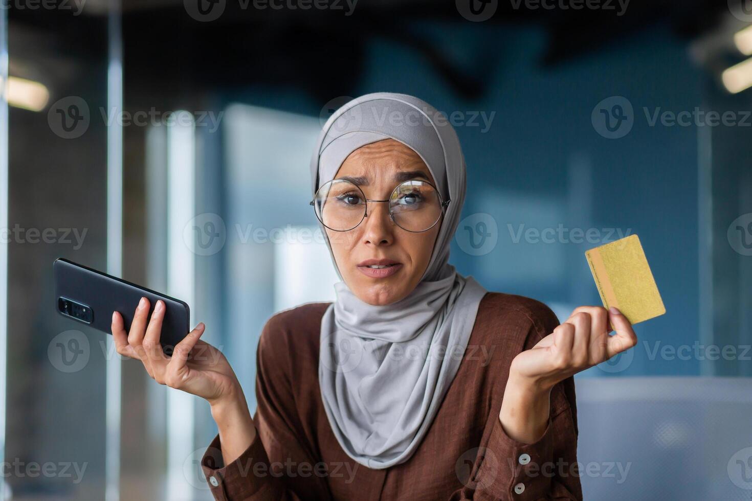 Close up photo. Portrait of a young Arab woman sitting in the office and holding a credit card in her hands. Problems with the account, money. He looks worriedly at the camera, spreads his hands. photo