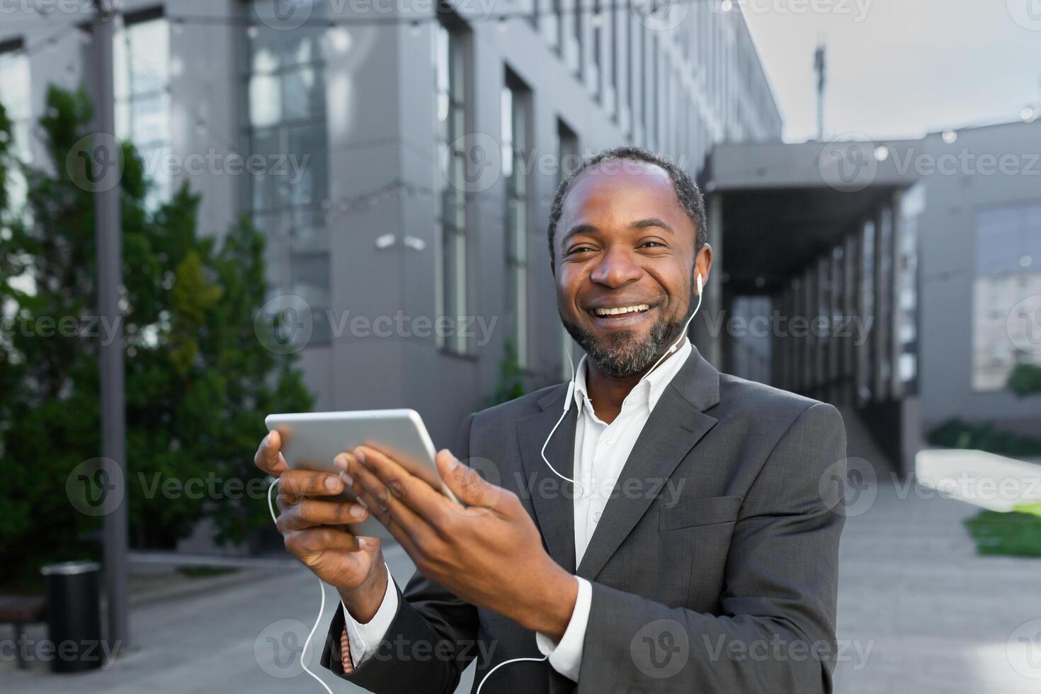 Portrait of young african american student in suit standing near campus wearing headphones. He holds a tablet in his hands, talks on a call, listens to music, looks at the camera, smiles. photo