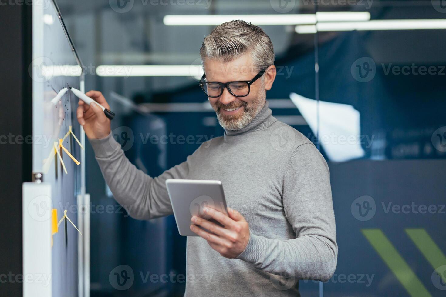 Mature man inside office near whiteboard with colored notes writing down strategy and business plan, gray haired businessman with tablet thinking at work inside office smiling happy with result. photo