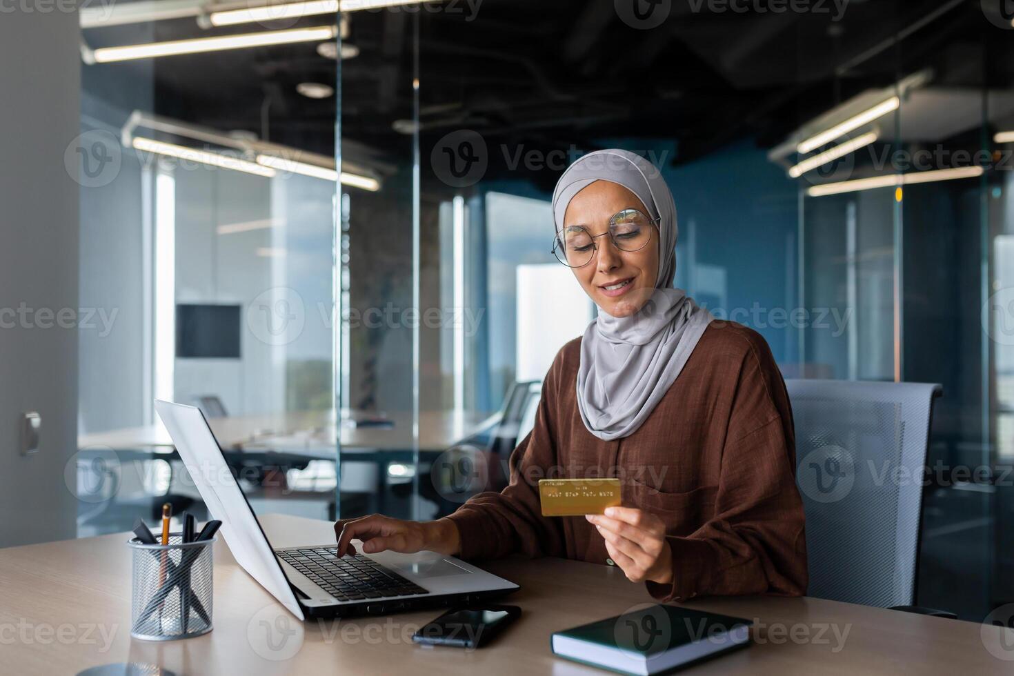 A young Muslim woman in a hijab is sitting in the office at a table with a laptop, holding and using a credit card. Pays bills, checks, online shopping. photo