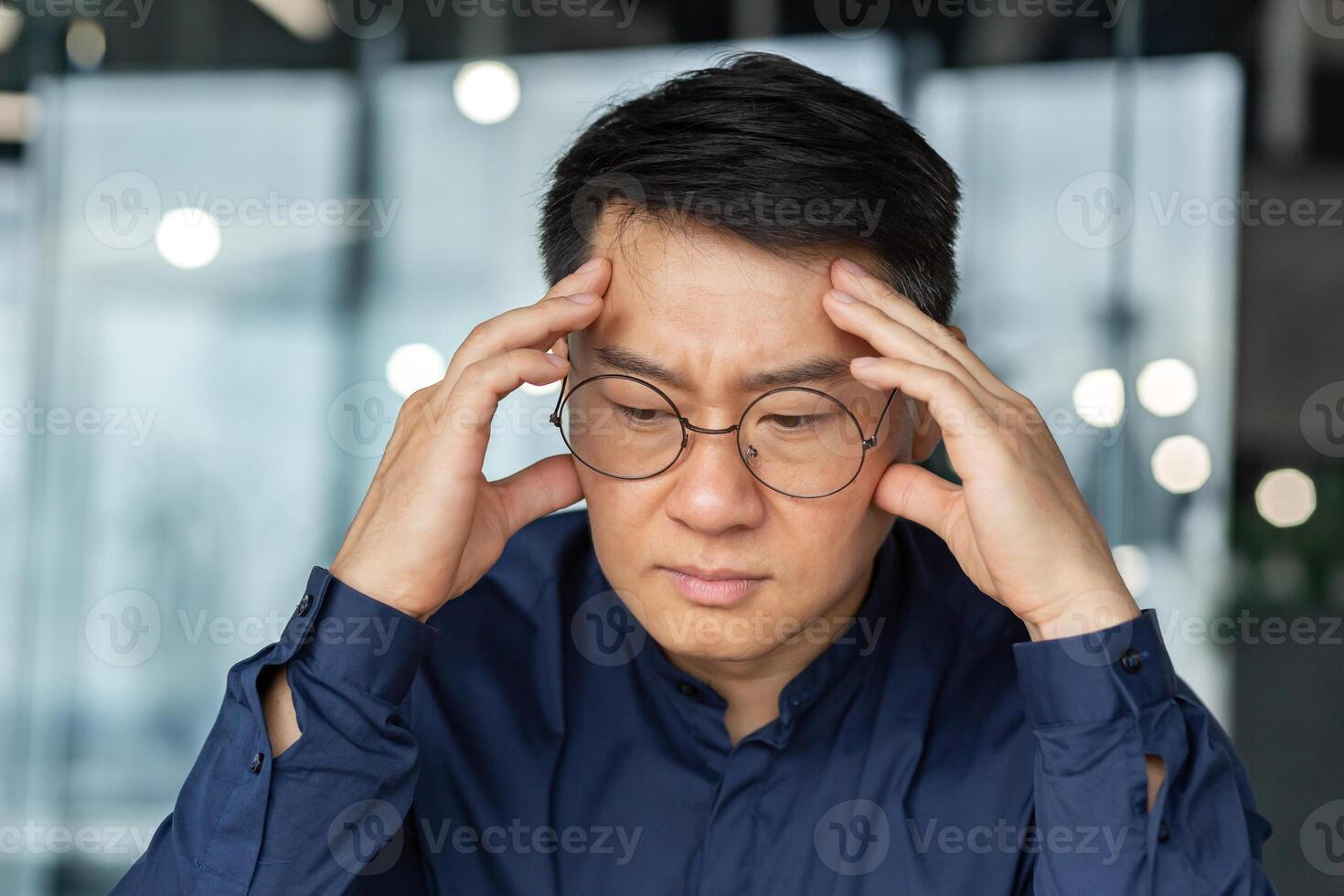 Closeup thinking businessman, Asian man in glasses and shirt holding hands on head making important decision, male manager working inside modern office building, thoughtful and serious boss. photo