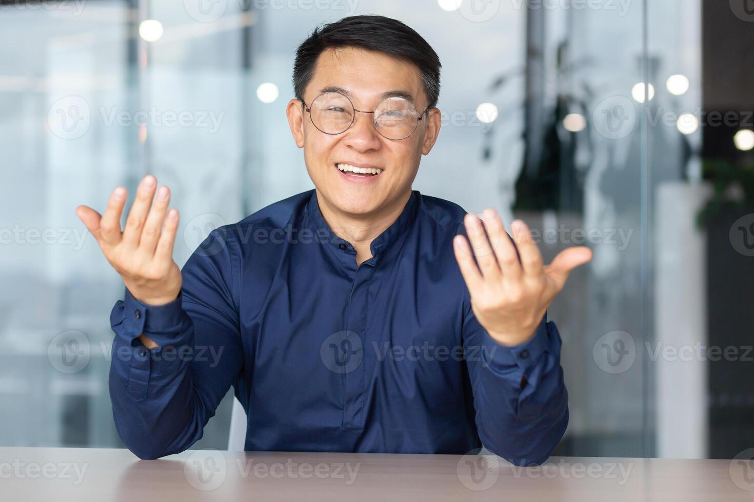 Portrait young male Asian teacher is lecturing students, taking exams online call. Sitting in the office at the table, smiling at the camera, explains, waving his hands. photo