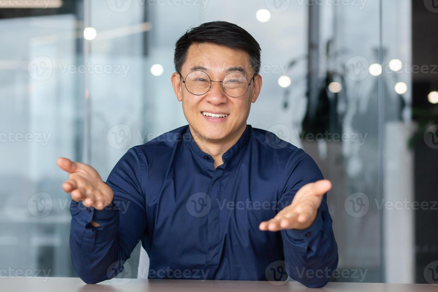 A handsome young Asian man is sitting in the office, looking at the camera, pointing at the camera, smiling. Blogger. online meeting. business training, teacher. photo