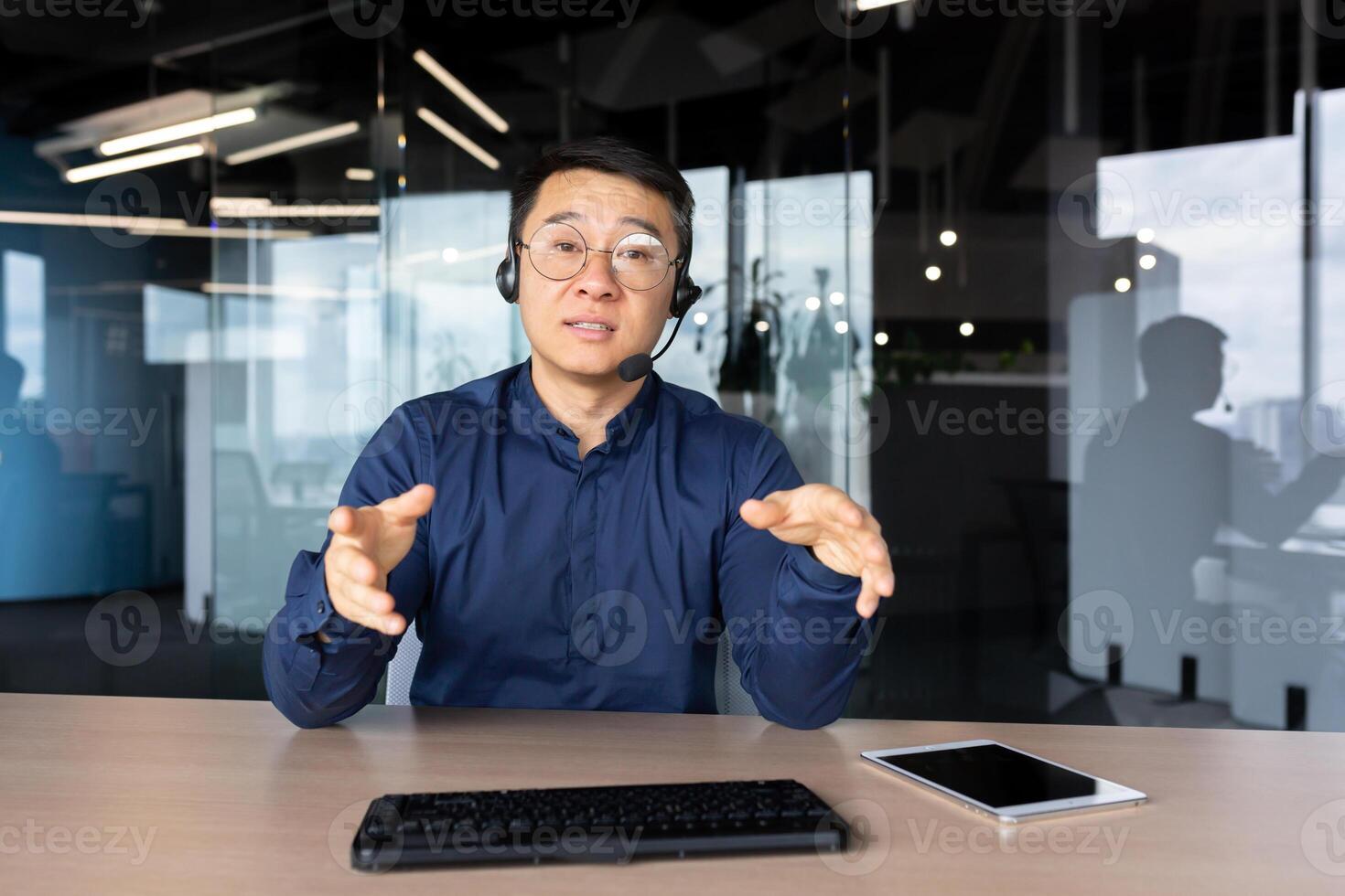 A young Asian man sits in the office at a desk in a headset, looks and talks to the camera. Explains, advises, conducts training, online meeting. photo