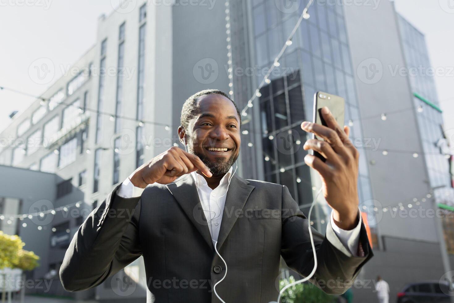 A senior black man in a suit is standing outside a business center and talking on the phone via call in headphones. photo