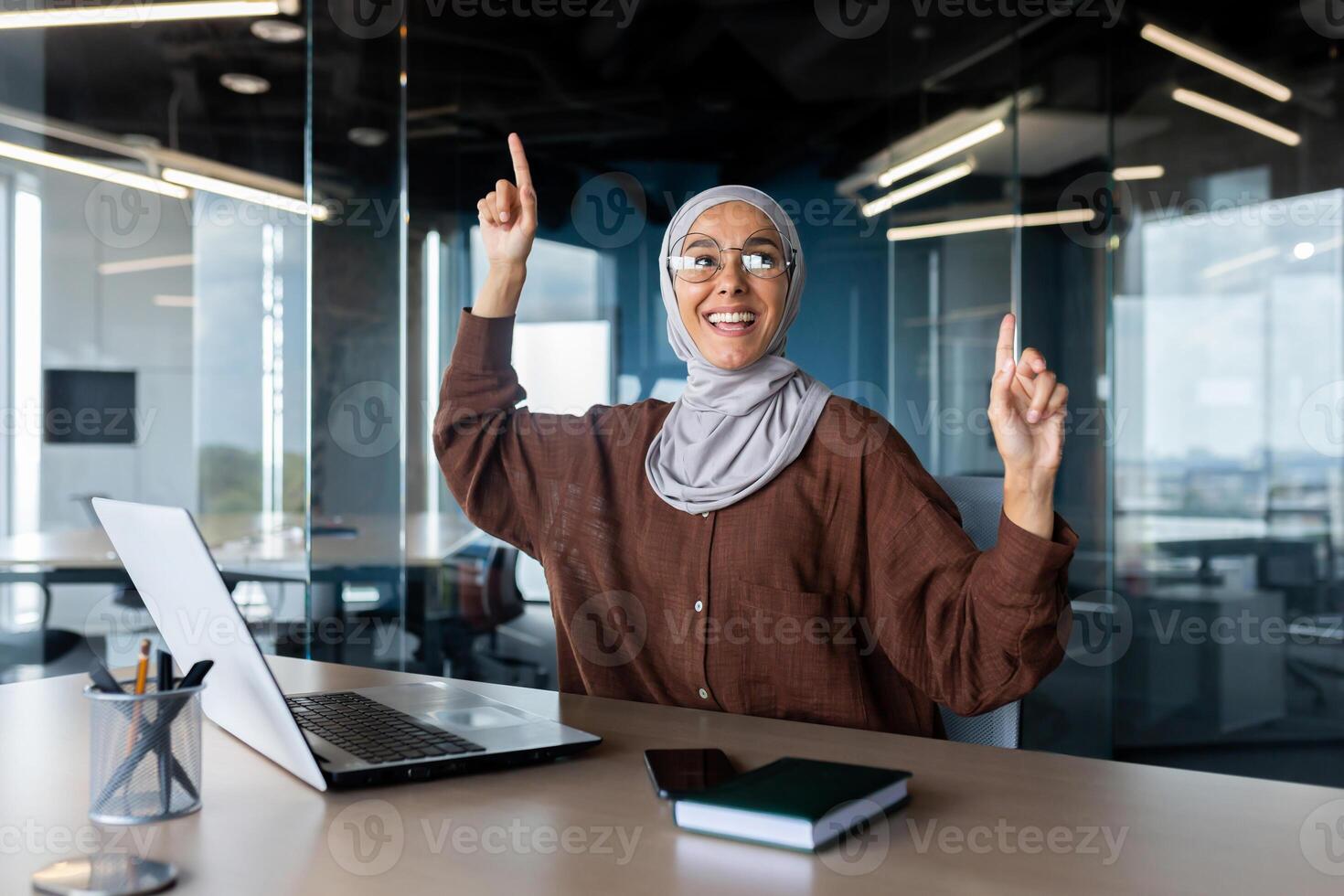 A young Muslim business woman in a hijab sits in the office at the table and dances. She looks happily at the camera, raised her hands up, celebrates success, a deal, a break. photo