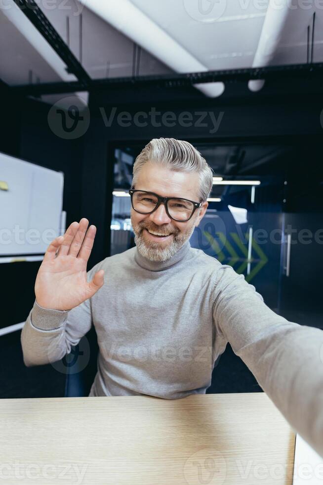Mature man inside office talking on call using app on phone, senior businessman having fun talking with friends and colleagues, taking selfie photo. photo