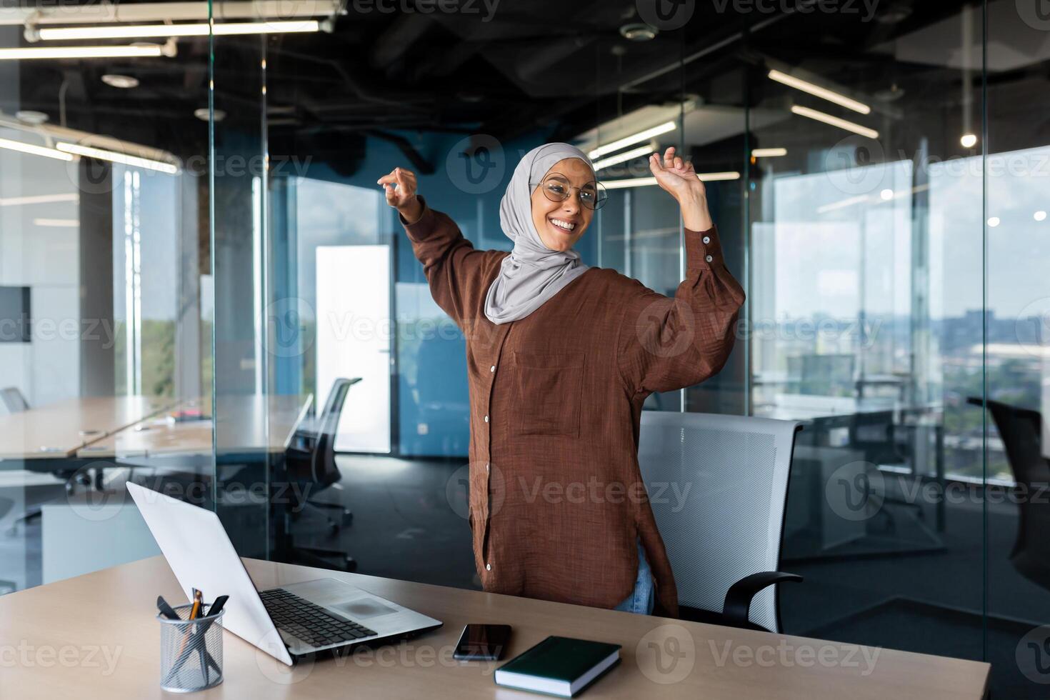 Young Arab business woman in hijab dances in the office near the table, listens to music on a laptop. Relaxed and smiling, she raised her hands up. Rest, break. photo