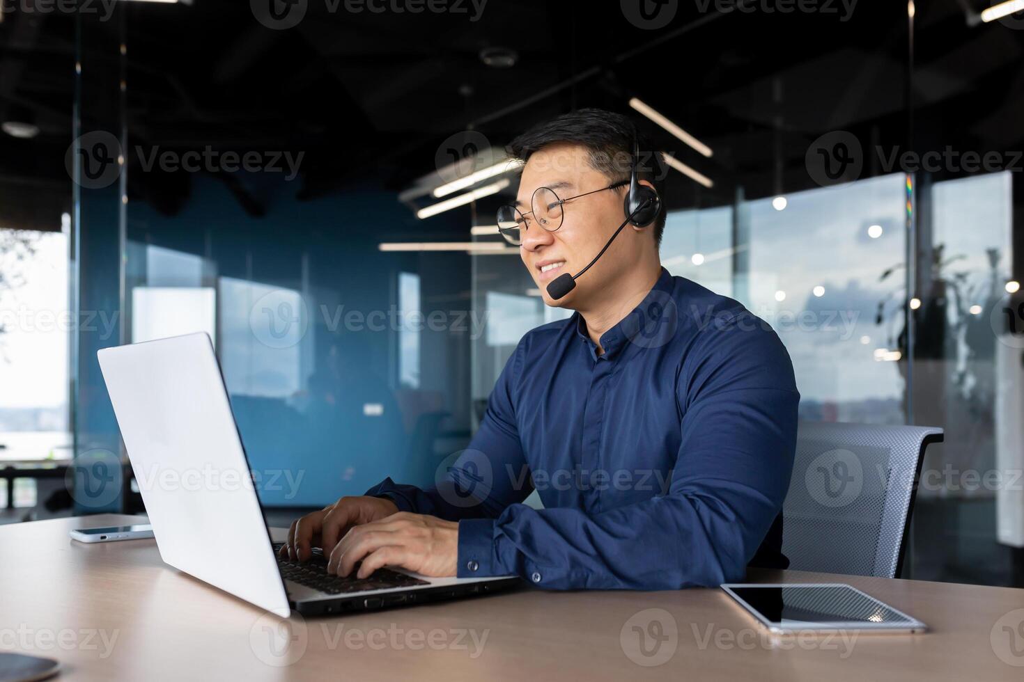 Young male Asian teacher is lecturing students, taking exams online via laptop call. Sitting in the office at the table in a headset, smiling at the camera. photo