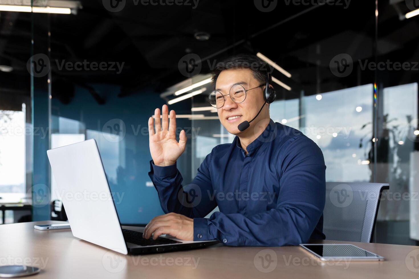 Successful smiling man working inside office using laptop and headset for call, asian cheerfully consulting tech support customer, businessman at work in shirt and glasses. photo