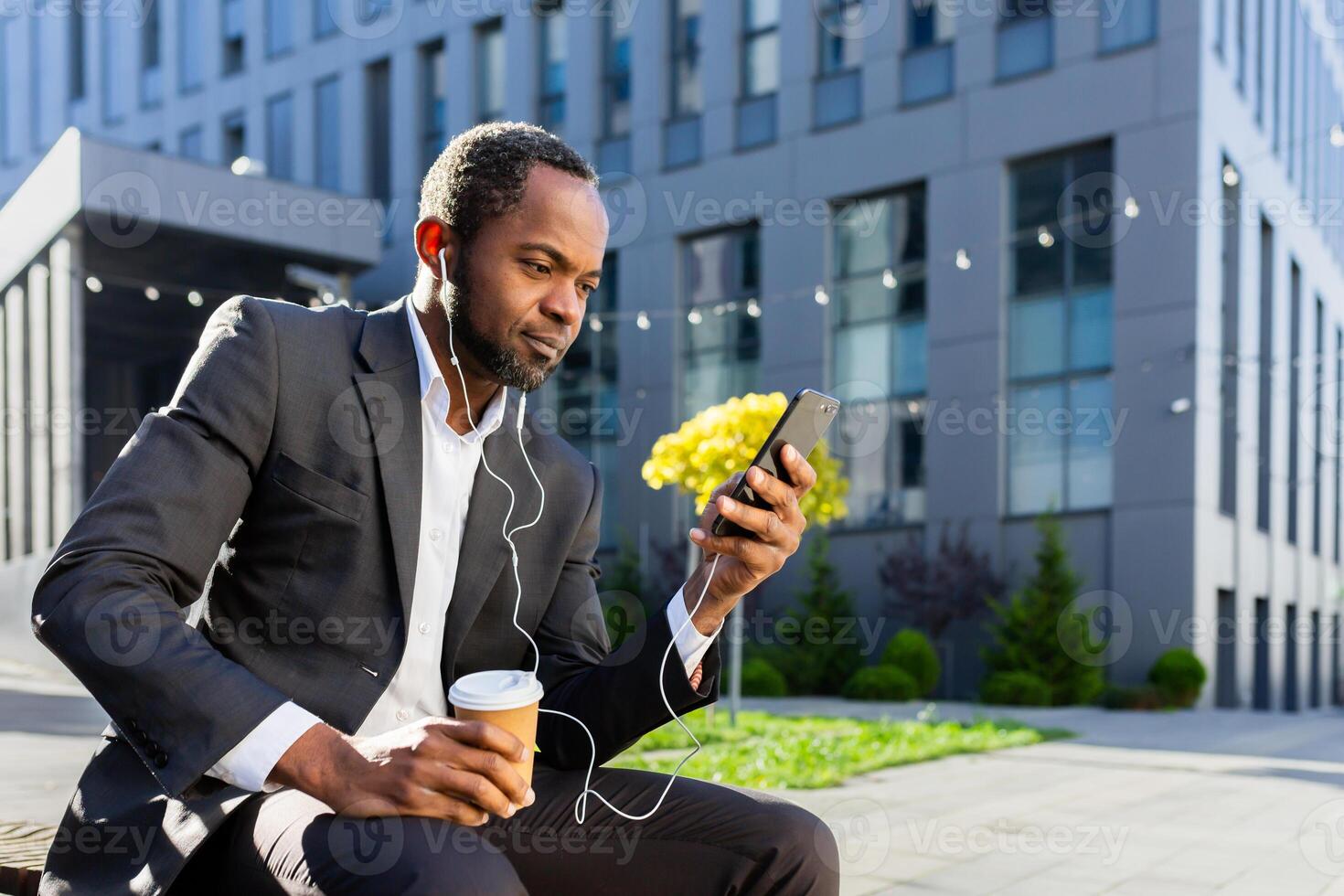 African American businessman sitting outside on a bench near an office center wearing headphones, holding a phone, a cup of coffee. Takes a coffee break, watches videos, listens to music, calls. photo