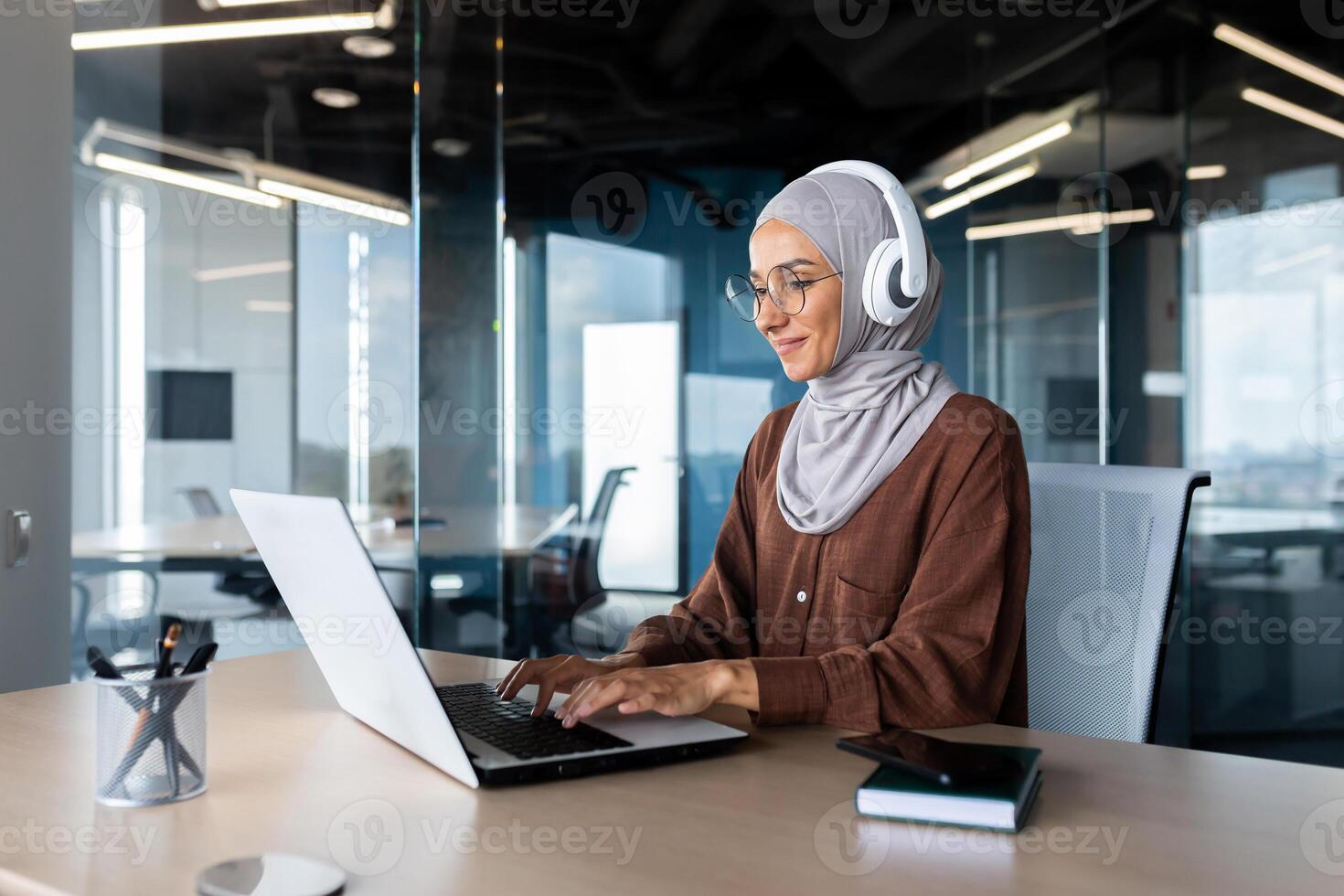 Online training. Young female student in hijab and headphones, sitting in the office at the table, studying remotely with a laptop. Typing on the keyboard.smiling. photo