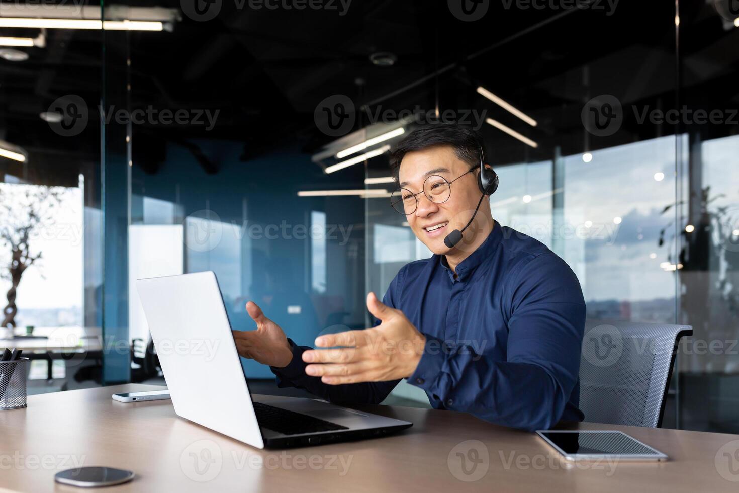 Asian with a headset is talking on call, man inside office smiling and gesturing joyfully in an online meeting, a businessman is telling his colleagues, a customer service tech support worker. photo