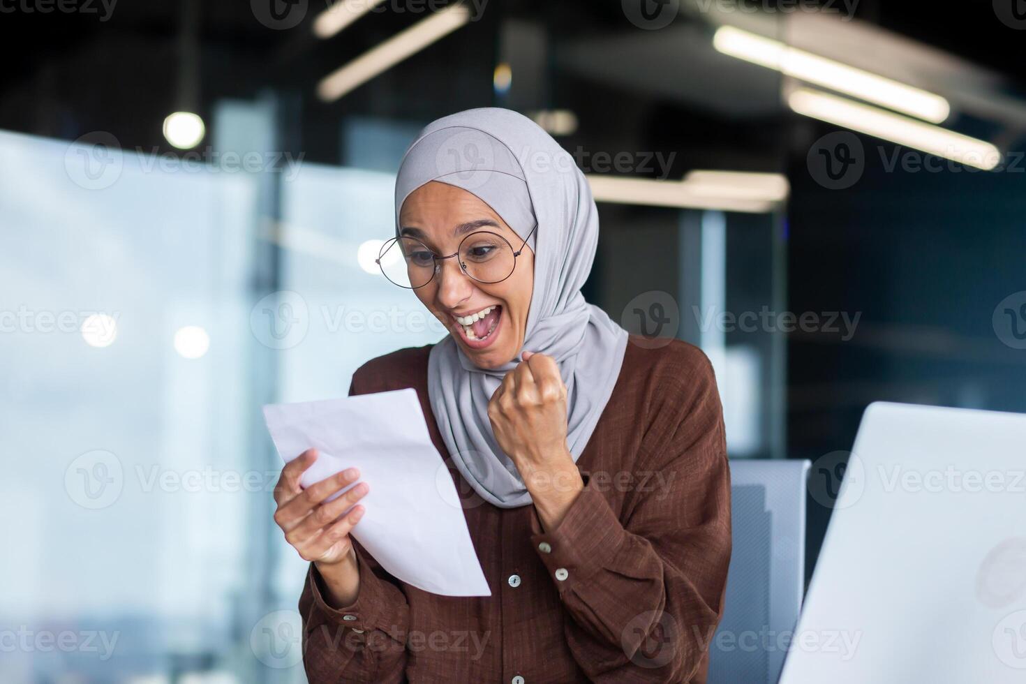 Young happy female student in hijab. Sitting at the table, holds a letter, received a notification of admission to the university, exam results. Celebrates, screams, shows success with his hand. photo