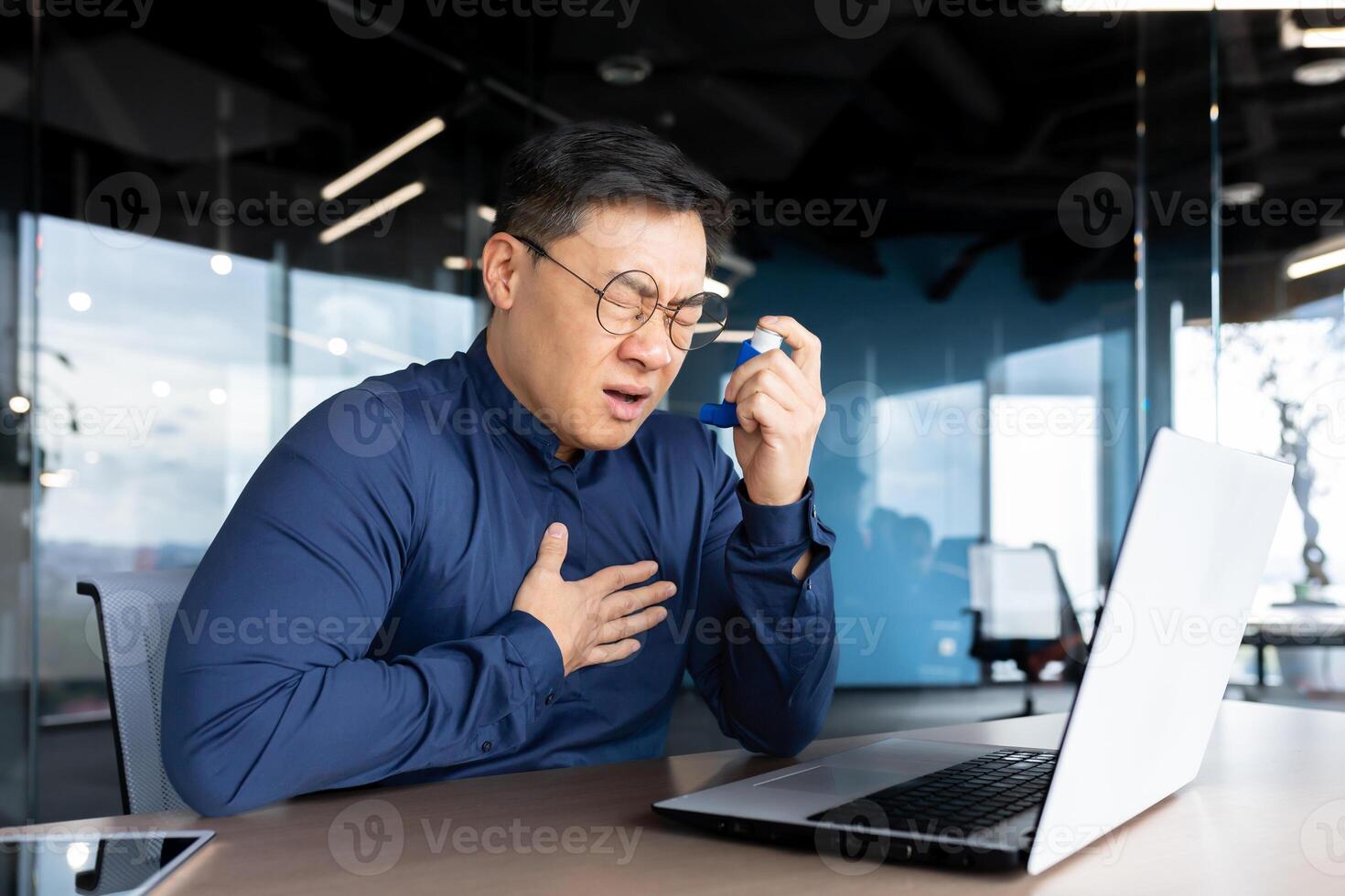 Young Asian man having asthma attack, panic attack, pneumonia at workplace. Sitting in the office at the table, holding his hands to his chest and an inhaler, he feels severe pain and is suffocating. photo