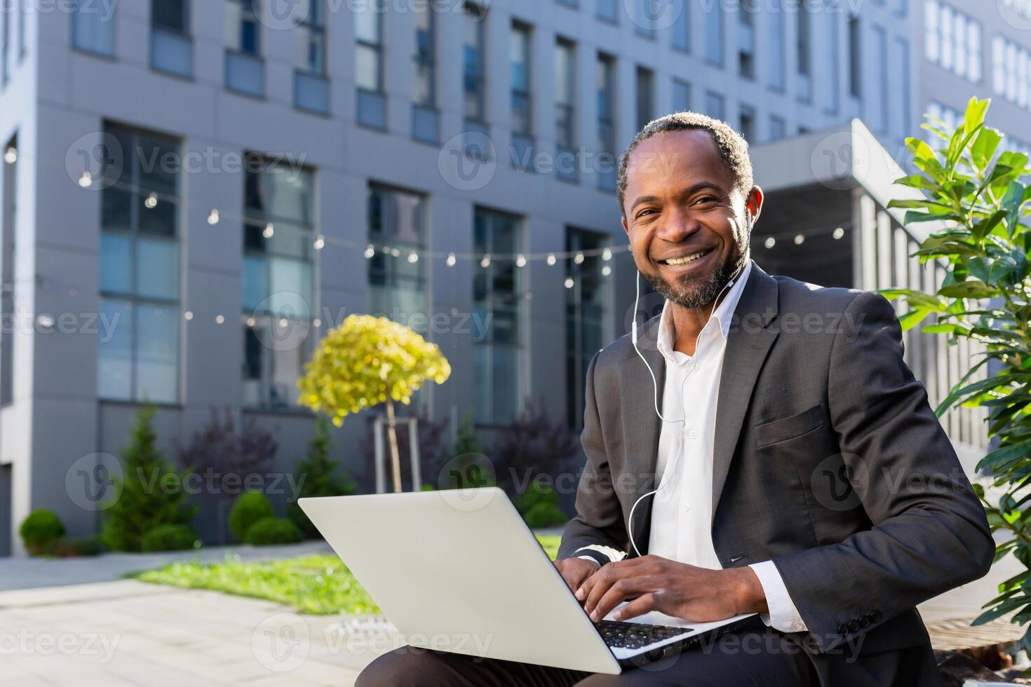 African american man in suit sitting near office center outside with laptop and headphones, and having online interview, talking on call. He looks at the camera, smiles. photo