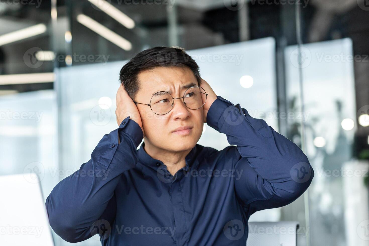 Noise in the office, Asian worker businessman covers his ears with hands from noise, man works inside office building complains about loud sound. photo