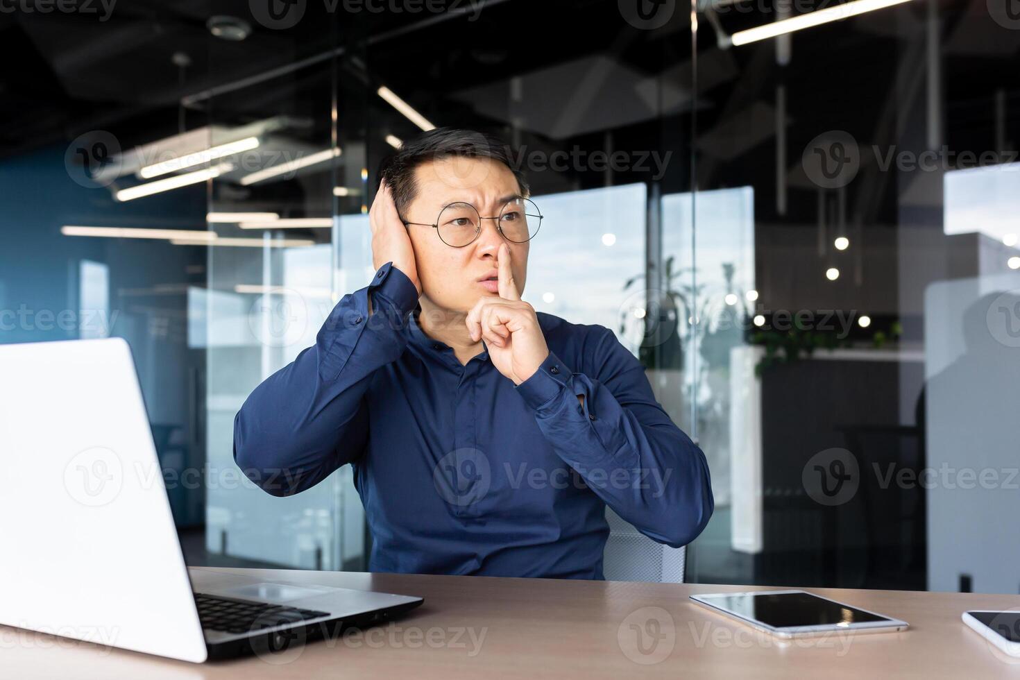 Upset and nervous asian man at workplace demanding silence from colleagues, businessman holding finger near lips and quarreling with co-workers, row inside office at workplace, discomfort at work. photo