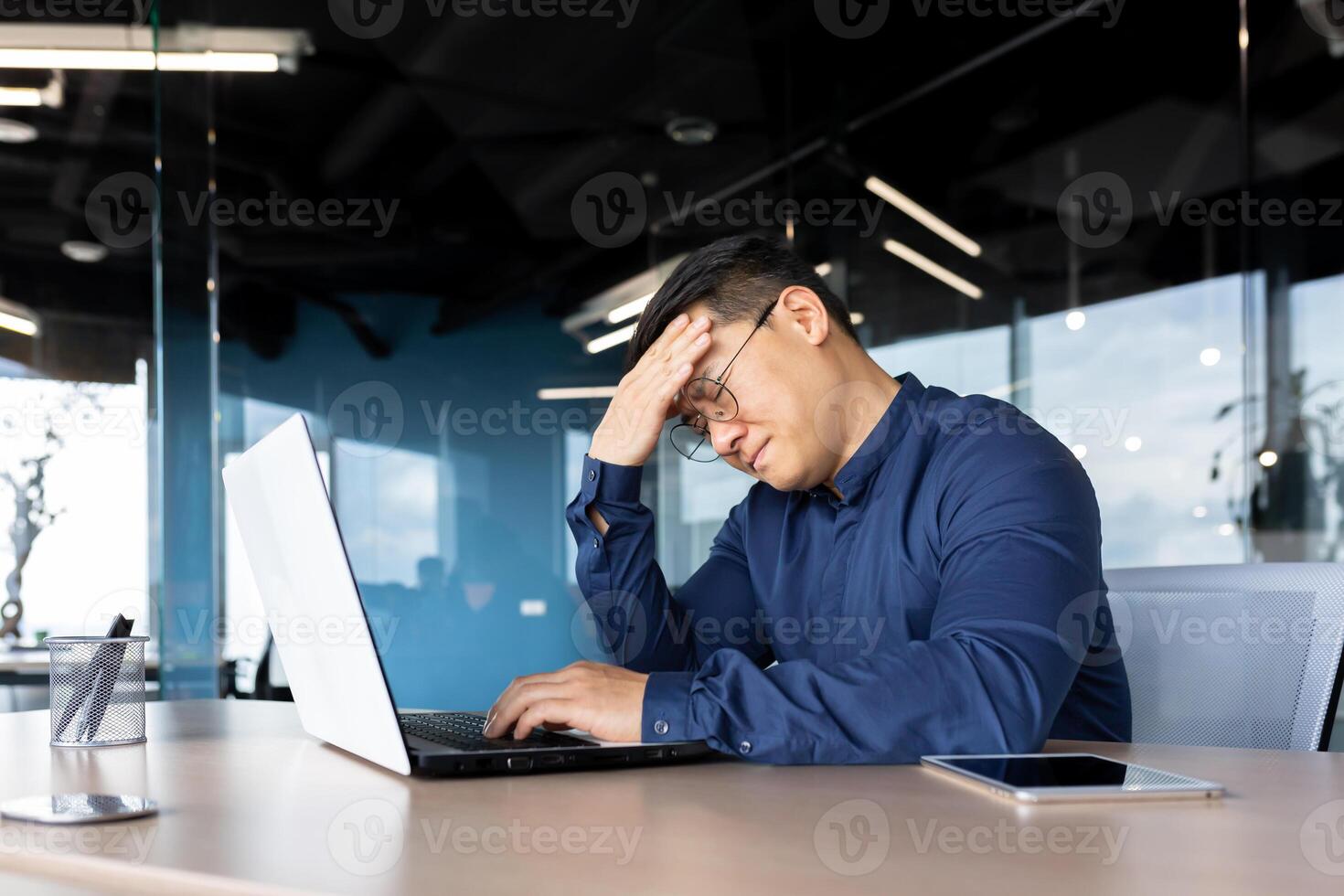A tired young Asian man sits in the office at the table, holds his head, feels a headache, tension, migraine after a hard overtime day, shakes. photo