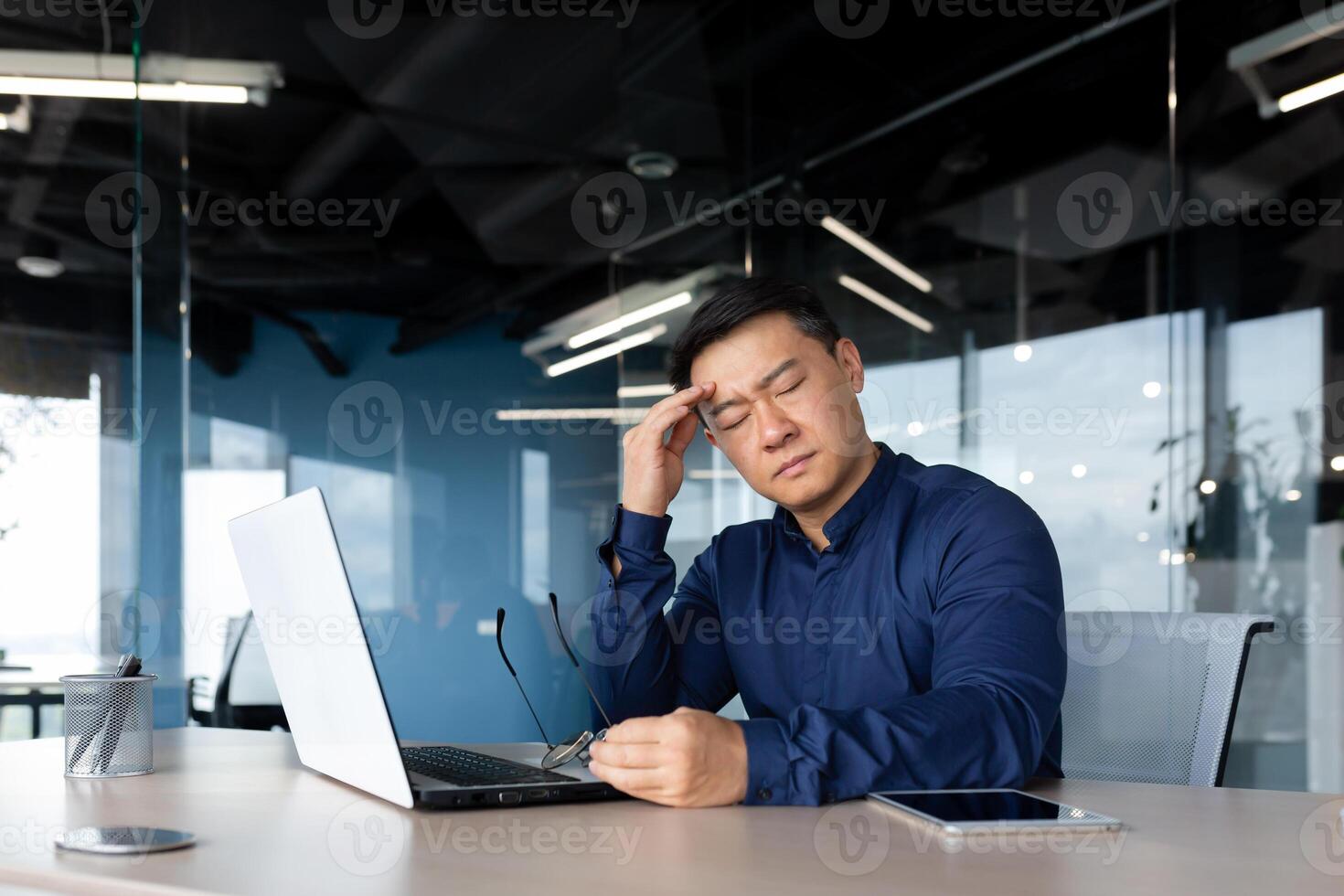 thinking sad asian man with closed eyes working in office indoors, businessman sitting at desk overworked and frustrated, mature man upset with achievement results. photo