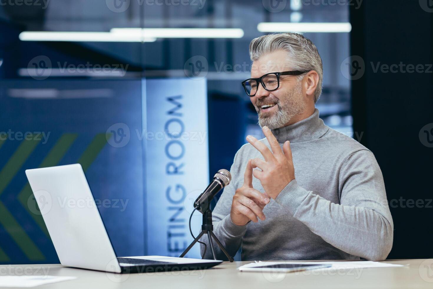 Senior gray-haired man sitting at a desk in an office with a microphone and laptop. Talks on a call. Conducts interviews, business meetings, webinars, online meetings. Shows, counts on fingers. photo