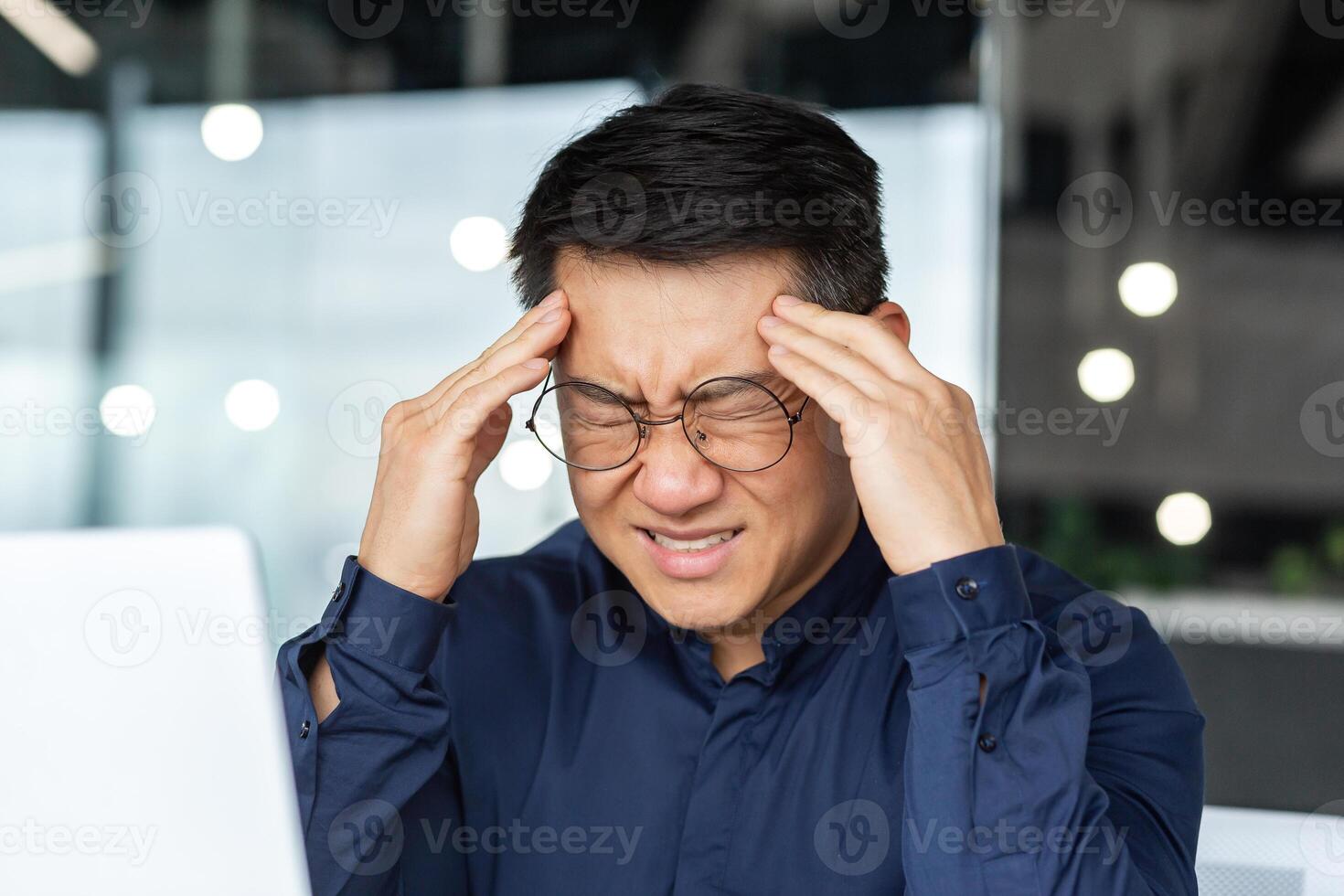 Close-up photo of sick Asian businessman having severe headache, male boss in glasses working inside modern office building, holding hands on head massaging, overtired at work.