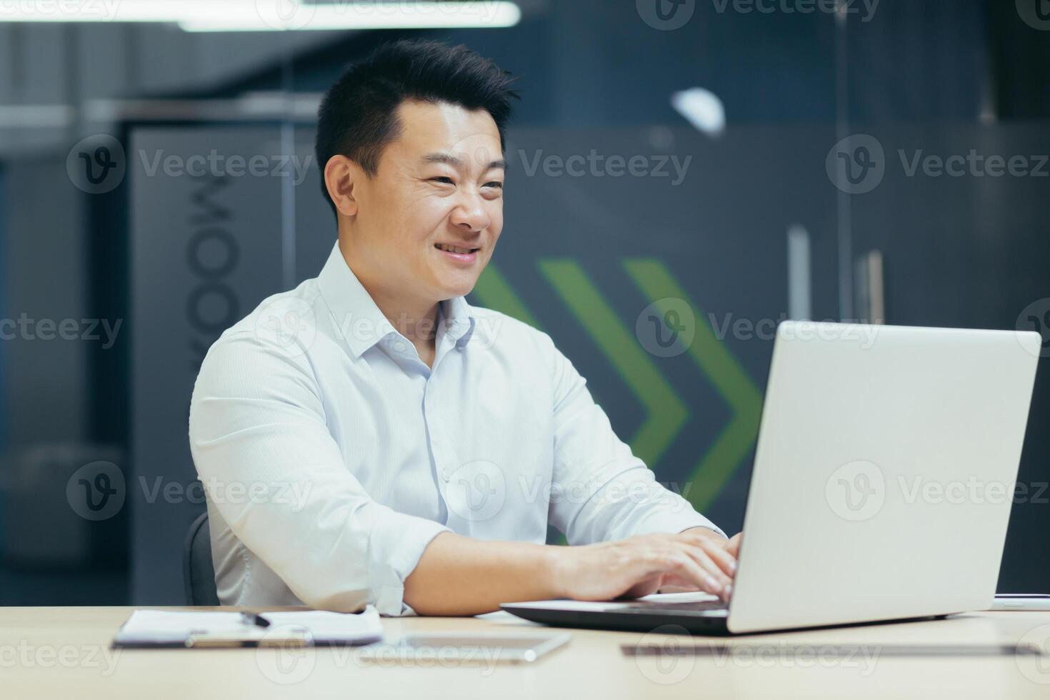 Portrait of a young smiling Asian businessman working in the office, sitting at a table, typing on a laptop. photo