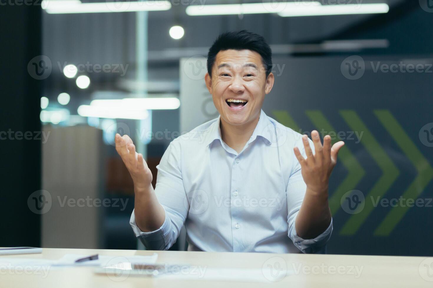 Portrait of a young happy Asian man sitting at an office desk in front of a camera and talking on a call. Gesturing with his hands, smiling at the camera. photo